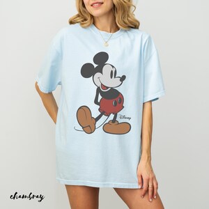 May include: Light blue t-shirt featuring a classic Mickey Mouse graphic. The cartoon character is depicted in his iconic pose, wearing red shorts and brown shoes. The Disney logo is visible. The word "chambray" is written in cursive at the bottom.