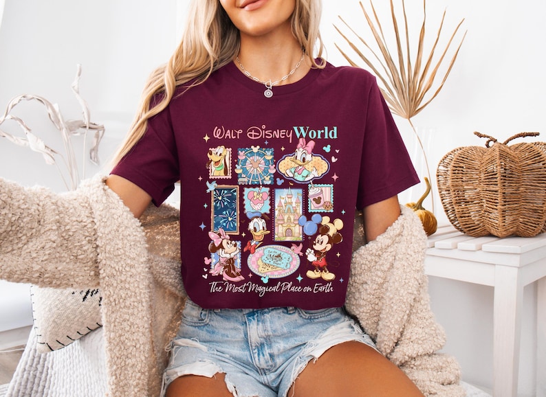 K&ouml;nnte beinhalten: Burgunderfarbenes T-Shirt mit einer farbenfrohen Grafik von Disney-Figuren und -Attraktionen, darunter Micky Maus, Donald Duck und Aschenputtels Schloss. Der Text "Walt Disney World" und "The Most Magical Place on Earth" sind ebenfalls abgebildet.