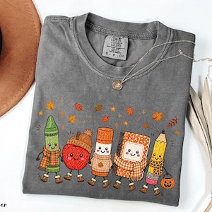 May include: A charcoal gray short-sleeve t-shirt featuring a graphic of five anthropomorphic school supplies including a green crayon, a red apple, a glue stick, a notebook, and a yellow pencil, all wearing autumn accessories and surrounded by falling leaves.