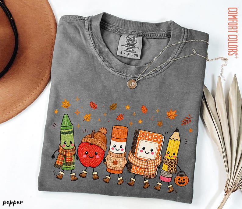 May include: A charcoal gray short-sleeve t-shirt featuring a graphic of five anthropomorphic school supplies including a green crayon, a red apple, a glue stick, a notebook, and a yellow pencil, all wearing autumn accessories and surrounded by falling leaves.