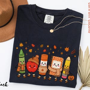 May include: A black crewneck t-shirt featuring a graphic of five anthropomorphic school supplies including a green crayon, an apple with an acorn hat, a glue stick, a notebook, and a yellow pencil, all wearing scarves and boots under autumn leaves.