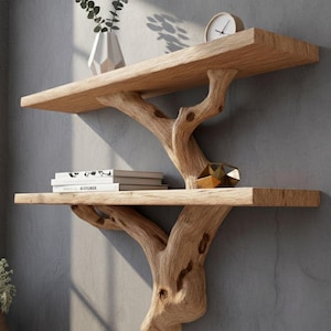 Puede incluir: Estantería de madera de dos niveles con una estructura de soporte natural en forma de rama de árbol. Los estantes son de madera clara y están adornados con objetos decorativos, como libros, un jarrón con vegetación y un pequeño reloj. La unidad está montada en una pared gris.