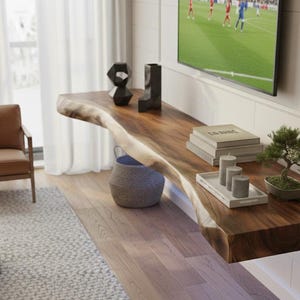 Pode incluir: Consola flutuante de madeira escura com borda natural. Apresenta livros, velas e um bonsai. A consola está fixada a uma parede branca, criando um ambiente moderno e elegante. Ideal para decorar salas de estar.