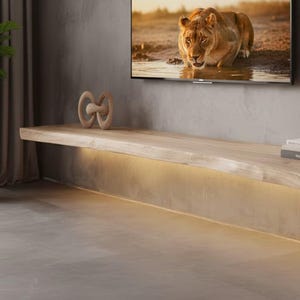 Puede incluir: Un salón moderno con una estantería flotante de madera clara con iluminación integrada. Un televisor muestra una escena de vida silvestre. Una planta en maceta y objetos decorativos completan la decoración minimalista. La habitación tiene una paleta de colores neutros.