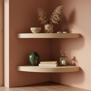 May include: Two light-coloured wooden shelves in a corner, displaying decorative items. The top shelf holds a woven vase with dried plants, a patterned bowl, and small stones. The bottom shelf features a green vase, books, and a candle lantern.
