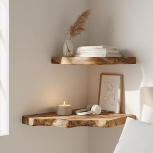 Puede incluir: Dos estanterías de esquina de madera con bordes naturales montadas en una pared blanca. La estantería superior contiene un jarrón con flores secas y libros. La estantería inferior muestra una vela encendida, auriculares y una impresión artística abstracta enmarcada.