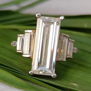 May include: A silver ring with a large, rectangular, clear gemstone as the focal point. The ring features smaller, rectangular gemstones on either side of the main stone. The ring is set on a green leaf.