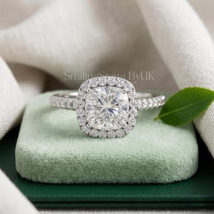 May include: A silver-toned ring featuring a large, cushion-cut center stone surrounded by a halo of small, round, clear stones. The band is also encrusted with small, round, clear stones. The text SmilingStone ByUK is visible above the ring.