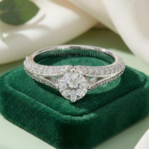 May include: A silver ring with a large, round, clear gemstone as the centerpiece. The band is encrusted with smaller, clear gemstones. The ring is set on a dark green velvet surface. The text "SmilingStone ByUk" is visible on the ring.