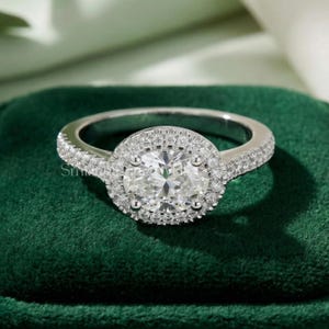 May include: A silver ring with a large, round, clear center stone surrounded by smaller stones. The band is also encrusted with small stones. The ring is set against a dark green velvet background.