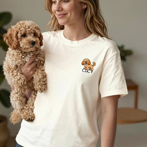 May include: A cream-coloured short-sleeve t-shirt featuring a small embroidered graphic on the left chest. The graphic depicts a light brown curly-haired puppy with the name Lucy written in black lettering directly underneath the dog illustration.