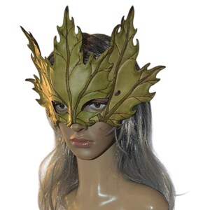 Op de afbeelding: Een handgemaakt, bladvormig masker in groen- en bruintinten. Het masker is gemaakt van leer en heeft ingewikkelde aderdetails. Het masker bedekt de ogen en neus, met een uitsparing voor de mond. Het masker wordt gedragen door een mannequin.