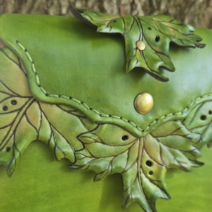 Puede incluir: Un bolso de cuero verde brillante con patrones de hojas en relieve, venas detalladas y pequeños recortes circulares. La solapa se asegura con un botón de presión metálico de color latón y los bordes tienen costuras visibles.