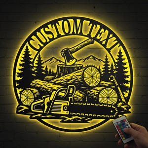 Op de afbeelding: Een cirkelvormig zwart metalen wandbord met achtergrondverlichting, met een bos, bergen, boomstammen, een bijl en een kettingzaag. De tekst "CUSTOM TEXT" staat in een boog bovenaan. Een afstandsbediening is op de voorgrond zichtbaar.