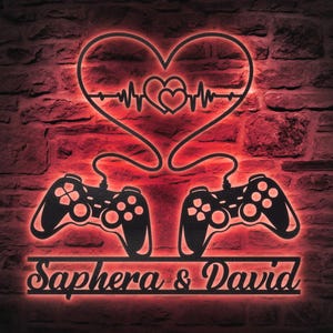 Puede incluir: Arte mural de metal negro con dos mandos de juego conectados a un diseño en forma de corazón con una línea de latidos. Los nombres "Saphera & David" se muestran debajo. El arte está iluminado con un resplandor rojo sobre un fondo de pared de ladrillo.