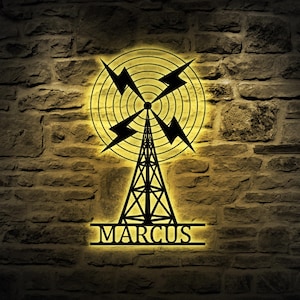 May include: Black metal wall art depicting a radio tower design with lightning bolts and concentric circles. The name "MARCUS" is displayed at the bottom. The art is illuminated with a warm yellow glow, mounted on a textured brick wall.