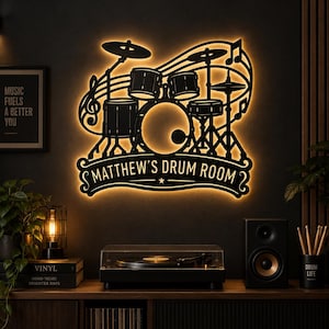Puede incluir: Un letrero de pared de metal negro con la silueta de una batería, notas musicales y el texto "MATTHEW'S DRUM ROOM" en una pancarta. El letrero está retroiluminado con un brillo cálido, creando un contorno distintivo tras el diseño de batería.