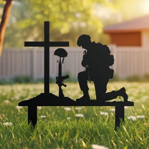 May include: Black metal silhouette of a kneeling soldier in front of a cross, helmet, and rifle. The memorial is set on a bench, placed in a grassy yard with a blurred background of a house and trees. A tribute to fallen soldiers.