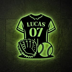 May include: A black wall-mounted sign featuring a glowing green outline of a baseball jersey with the name LUCAS and the number 07 printed on the front, accompanied by a baseball glove and a baseball, all illuminated with a bright green light.