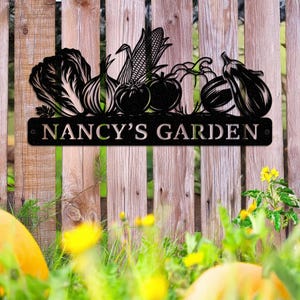 May include: Black metal garden sign featuring a variety of vegetables, including lettuce, onions, corn, tomatoes, and eggplant. The sign reads "NANCY'S GARDEN" in a decorative font, mounted on a wooden fence.