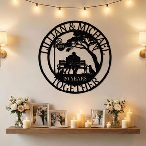 May include: A black metal wall art piece with the names "LILLIAN & MICHAEL" and "20 YEARS TOGETHER" encircling a silhouette of a couple on a bench, a dog, and a tree. The year "2006" is also included. The piece is displayed above a shelf with candles and framed photos.
