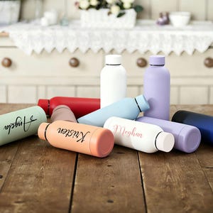 May include: A collection of insulated water bottles in various colors, including white, red, blue, and pastel shades. Some bottles are personalized with names like "Angela," "Kristen," and "Meghan." The bottles are displayed on a wooden surface.