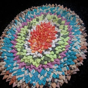Puede incluir: Una alfombra redonda hecha a mano, con un diseño multicolor. El centro presenta tela naranja, rodeada de capas de tela verde, azul, morada y estampada. La alfombra está sobre un fondo negro.