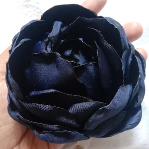 May include: A dark navy blue fabric flower with layered petals. The flower has a pearl-like bead attached to the bottom. The flower is held in a hand.