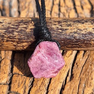 Peut inclure: Un collier pendentif en pierre précieuse rose. La pierre brute taillée et facettée est d'un rose vif et est suspendue à un cordon noir. Le collier est présenté sur un fond en bois.