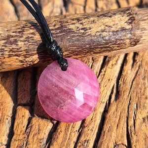 Puede incluir: Collar con colgante de piedra preciosa rosa, redondo y facetado. El colgante cuelga de un cordón negro. La piedra tiene una superficie pulida, sobre un fondo de madera texturizada.
