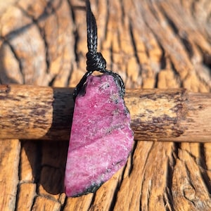 Peut inclure: Pendentif en pierre précieuse rose brute avec un cordon noir. Le pendentif a une forme brute et irrégulière, affichant une teinte rose vibrante avec des bords plus foncés. Le cordon est noué en haut, prêt à être porté. Fond en bois.