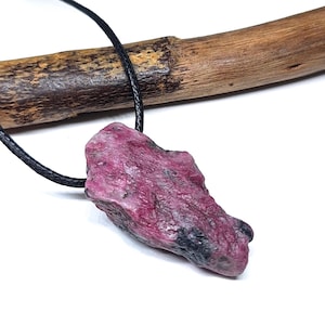 May include: A raw, unpolished magenta and dark gray stone pendant suspended from a thin black cord. The stone has an irregular, jagged shape with a rough, textured surface, showcasing natural color variations throughout the mineral structure.