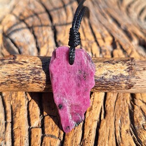 Peut inclure: Un pendentif en rubis rose brut avec un collier en cordon noir. La pierre précieuse a une texture naturelle rugueuse et est suspendue à un cordon noué. Le fond est une surface en bois texturé.