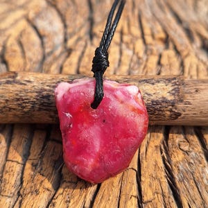 Op de afbeelding: Een ruwe, rozerode edelsteen hanger met een zwart koord. De onregelmatig gevormde steen heeft een natuurlijk, organisch uiterlijk. De hanger hangt tegen een achtergrond van getextureerd hout.