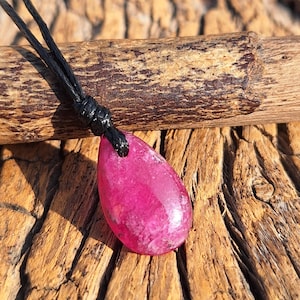 May include: A magenta teardrop pendant necklace. The polished stone hangs from a black cord. The necklace is displayed on a wooden surface with a natural grain.