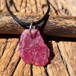May include: A raw, deep pink gemstone pendant with a rough, textured surface and irregular edges. The stone is suspended from a thin, black cord by a simple silver-toned metal jump ring, creating a necklace with a natural, unpolished appearance.