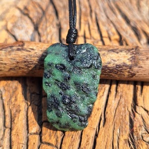 Peut inclure: Pendentif en émeraude brute sur cordon noir. L'émeraude est d'un vert profond avec des inclusions noires et une texture naturelle. Le pendentif est mis en valeur sur un fond en bois.