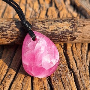 Puede incluir: Un colgante de piedra preciosa rosa facetada en forma de lágrima, suspendido de un cordón negro. El colgante tiene una superficie texturizada y es de un color rosa brillante. El cordón está anudado en la parte superior. El colgante está sobre un fondo de madera.