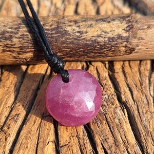 May include: A faceted, round, ruby-colored gemstone pendant necklace. The pendant is suspended from a black cord. The background is a wooden surface with a natural, rustic appearance. The pendant is the focal point.