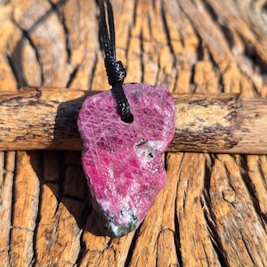 May include: A raw, pink ruby pendant with a black cord. The gemstone has a rough, textured surface and a hole drilled through the top for the cord. The pendant is displayed on a wooden background.