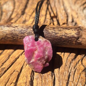 Peut inclure: Un pendentif en pierre brute rose non polie suspendu à un fin cordon noir. La pierre présente une forme irrégulière et dentelée avec une surface texturée et des teintes magenta profond, fixée au cordon par un petit trou percé au sommet.