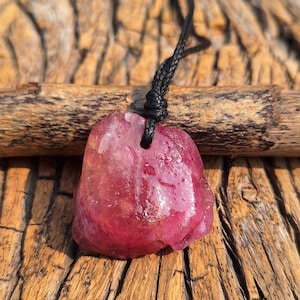 Puede incluir: Colgante de piedra preciosa rosa-roja en bruto, de forma irregular, suspendido de un cordón negro trenzado. El colgante se exhibe sobre un fondo de madera envejecida, resaltando su textura y color natural.