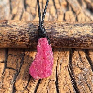 Könnte beinhalten: Ein Anhänger aus rohem, rosa Rubin an einer schwarzen Lederschnur. Der Rubin hat eine raue, natürliche Textur und eine unregelmäßige Form. Die Schnur ist oben geknotet, wodurch eine einfache, rustikale Halskette entsteht. Der Hintergrund ist eine Holzoberfläche.
