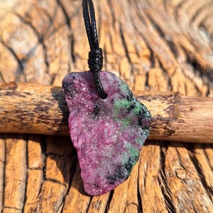 Peut inclure: Pendentif en rubis brut avec un cordon noir. Le rubis présente un mélange de couleurs rose, vert et noir. Le pendentif est de forme irrégulière, semblant être une pierre naturelle. Fond en bois.