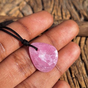 May include: A pink gemstone pendant, cut in a teardrop shape, suspended on a black cord. The pendant has a faceted surface, catching the light. The cord is knotted at the top. The background is a wooden surface.