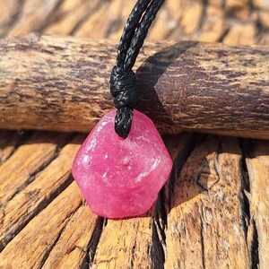 May include: A pendant featuring a raw, pentagonal-shaped pink gemstone suspended from a black cord. The stone has a translucent, crystalline texture with visible natural facets and irregular edges, securely knotted to the dark, braided cord.