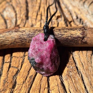 May include: A raw, deep pink ruby gemstone pendant suspended from a thin black cord. The stone features an irregular, natural shape with visible dark inclusions and a textured surface, showcasing its unpolished, organic appearance as a necklace.