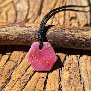 Op de afbeelding: Een ruwe, zeshoekige roze robijn hanger hangend aan een dun zwart koord. De edelsteen heeft een getextureerd, mat oppervlak met natuurlijke insluitingen en kleurvariaties, wat een levendige magentakleur door de hele steen laat zien.