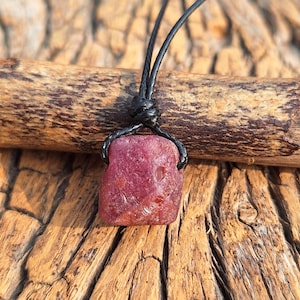 May include: A raw, reddish-pink gemstone pendant on a black cord. The irregularly shaped stone appears to be a natural crystal. The necklace is set against a backdrop of weathered wood, highlighting the pendant's colour and texture.