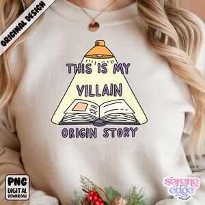 May include: A cream-colored sweatshirt featuring a cartoon design with the text "THIS IS MY VILLAIN ORIGIN STORY." The design includes an open book illuminated by a lamp. The words "ORIGINAL DESIGN" and "PNG DIGITAL DOWNLOAD" are also visible.